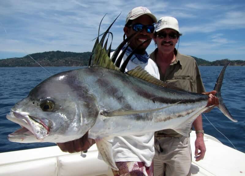 Two smiling anglers holding a Panama trophy roosterfish fish with Isla Ladrones in the background